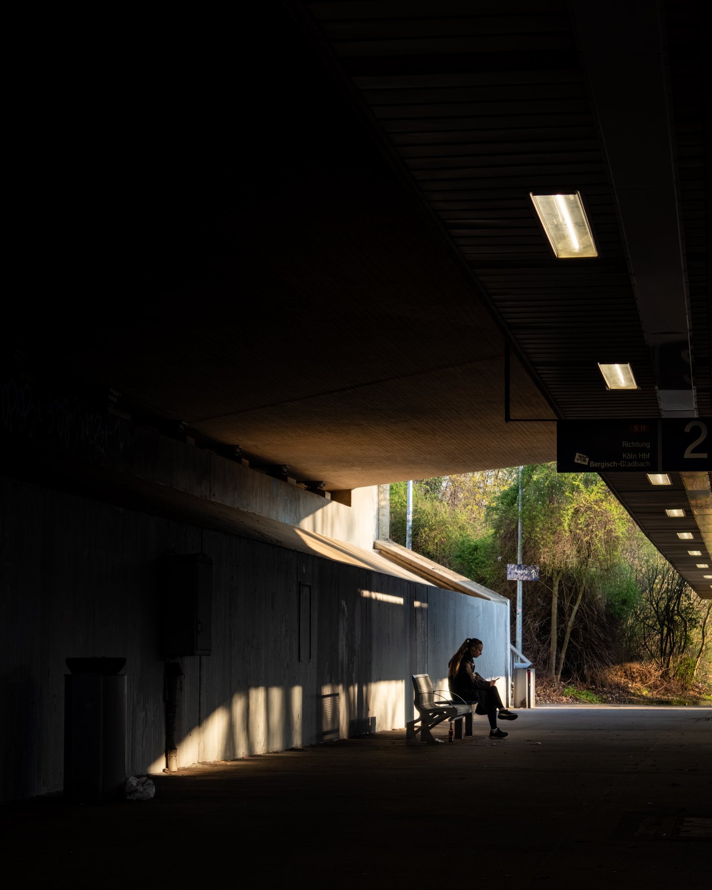 A figure sitting alone on a bench in a shaft of evening light under a train platform overhang