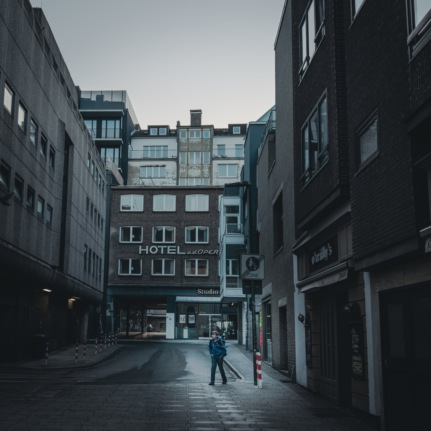 A person crossing an empty street framed by a hotel sign reading Hotel a.d. Oper at dusk