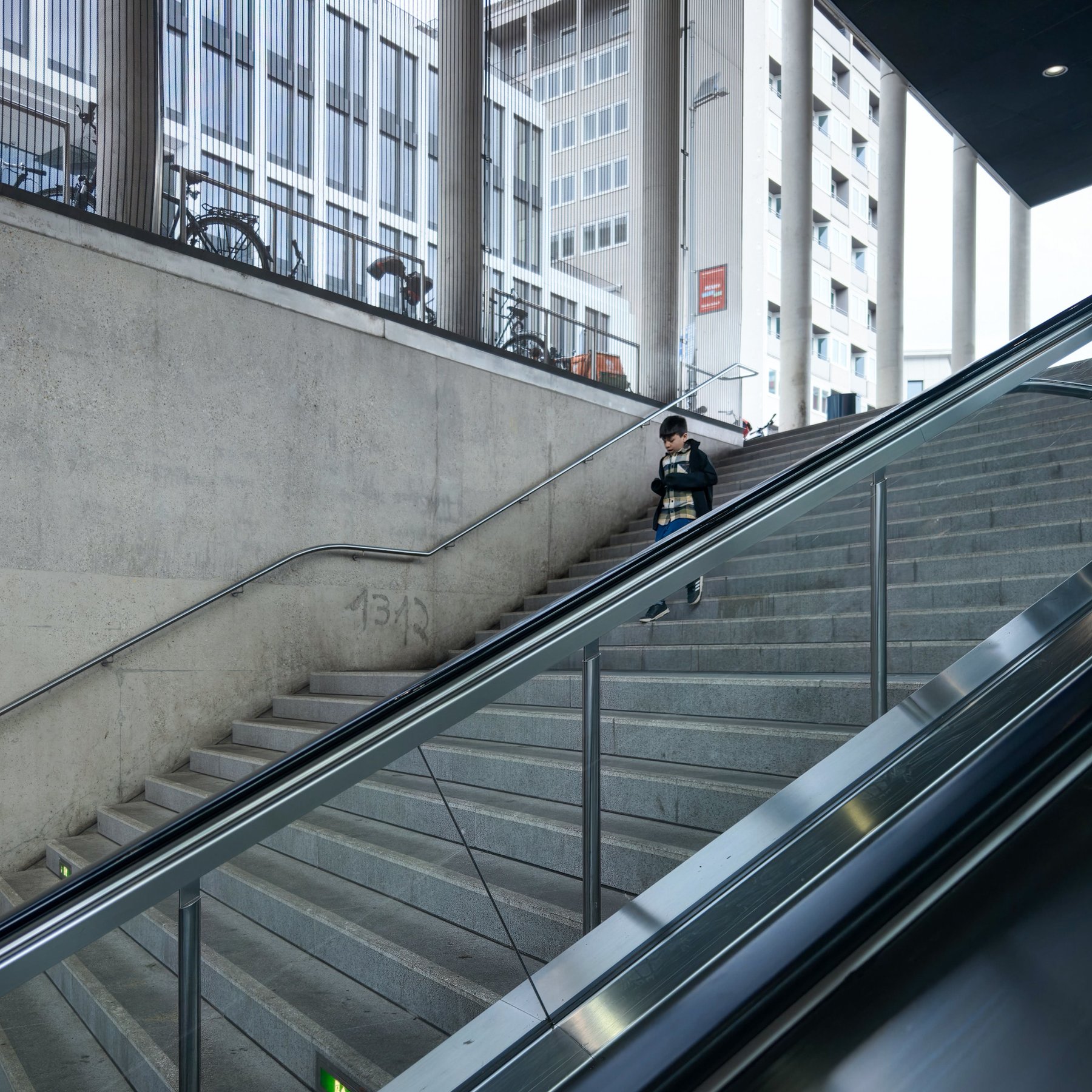 A boy descending a concrete staircase between glass railings, apartment buildings in the background